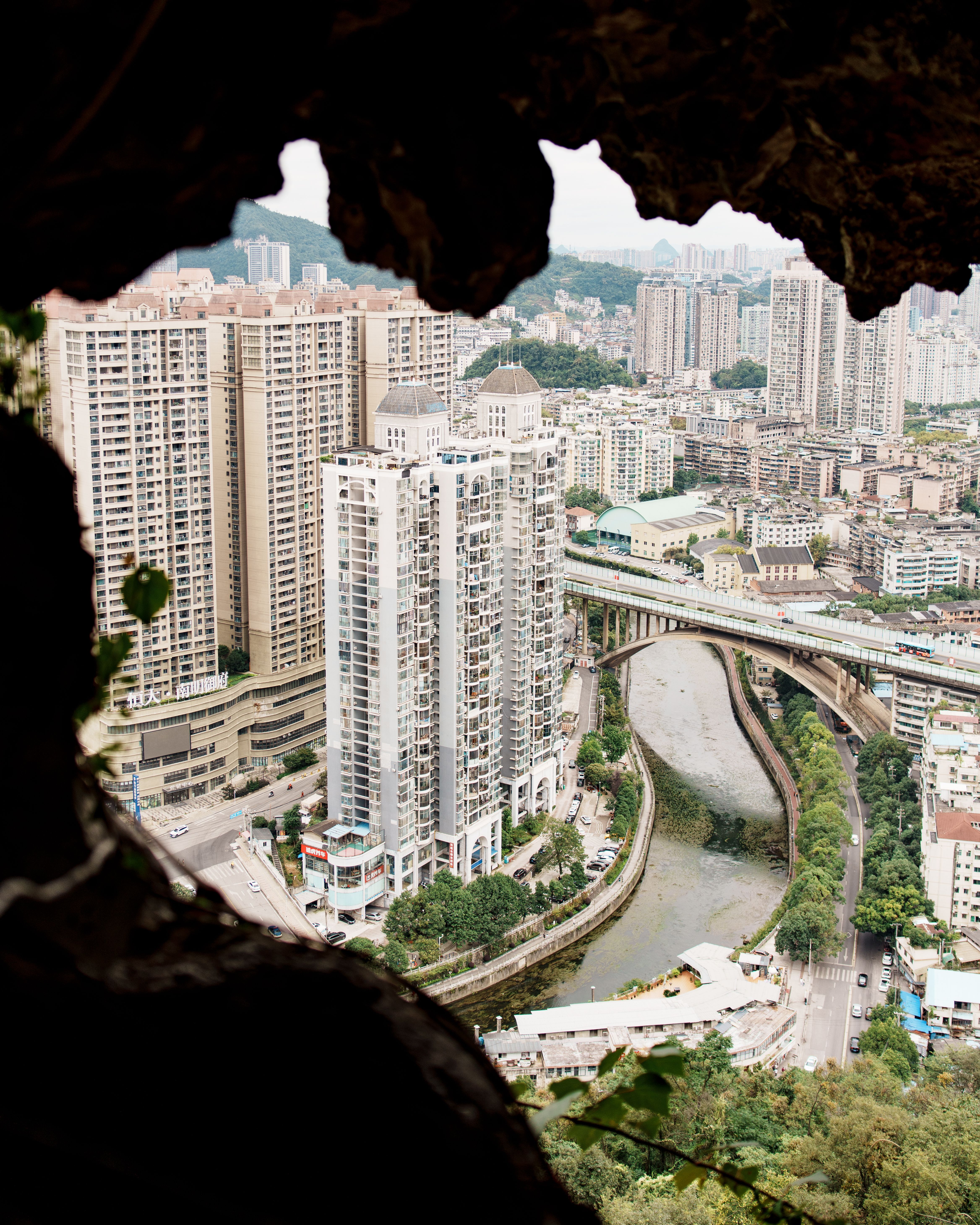 Unique view of Guiyang cityscape through a cave, showcasing modern high-rise buildings and a winding river.