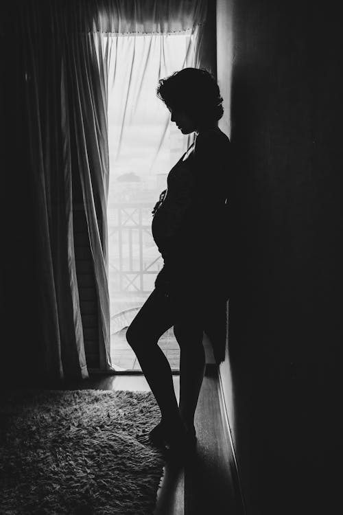 Silhouette of unrecognizable pregnant woman standing near window at home