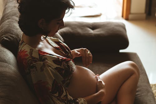 From above of crop young pregnant female in crop blouse relaxing on comfortable couch and touching belly gently at home
