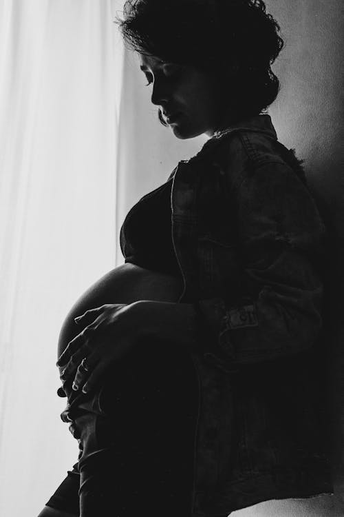 Free Side view of black and white crop calm pregnant woman in crop top and jacket standing near wall at home and touching tummy Stock Photo