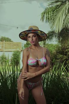Woman in vibrant bikini and hat poses amid palm fronds in Argentina garden setting.
