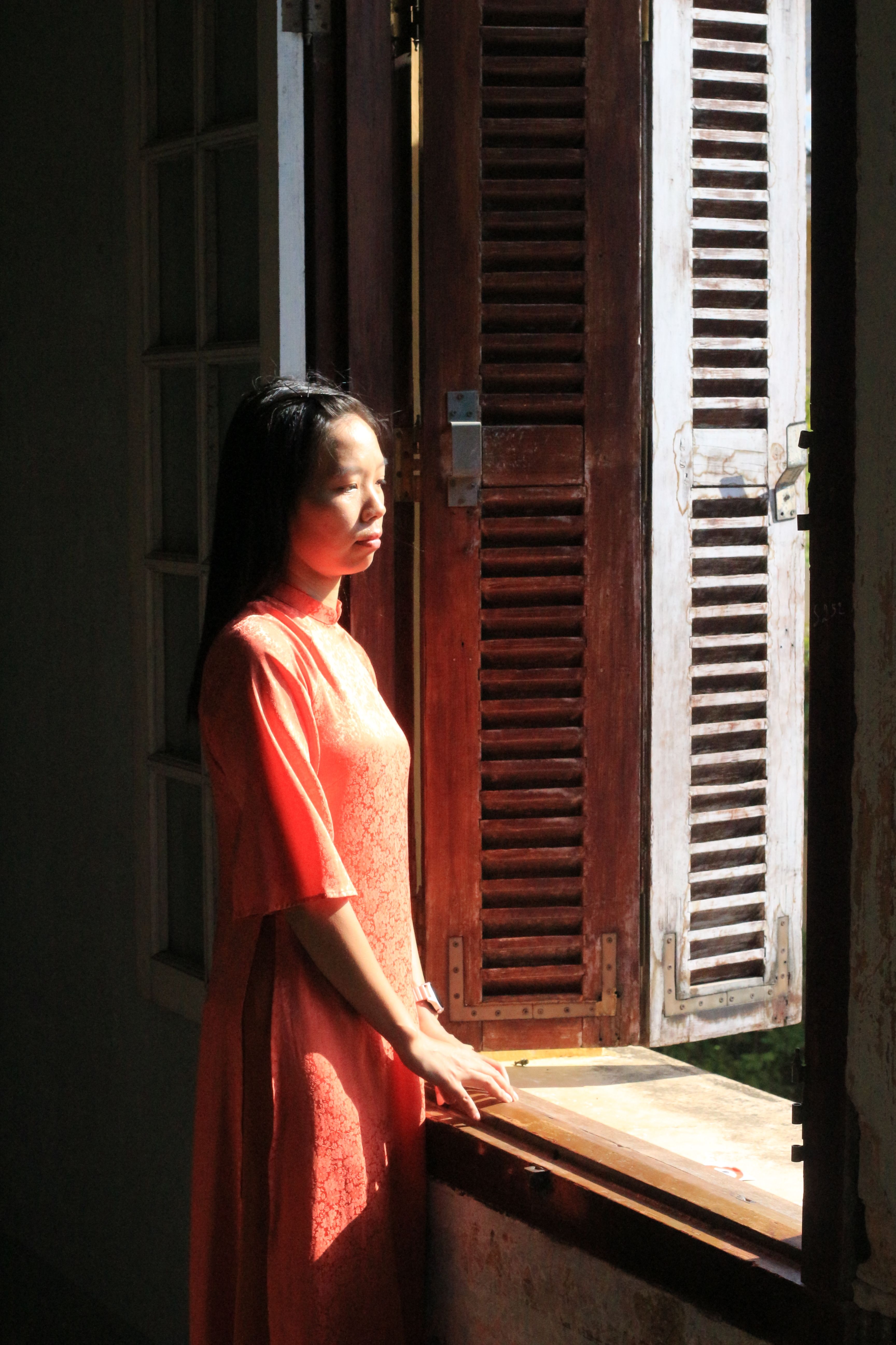 A woman in ao dai stands by a sunlit window, capturing a peaceful moment in Hue, Vietnam.