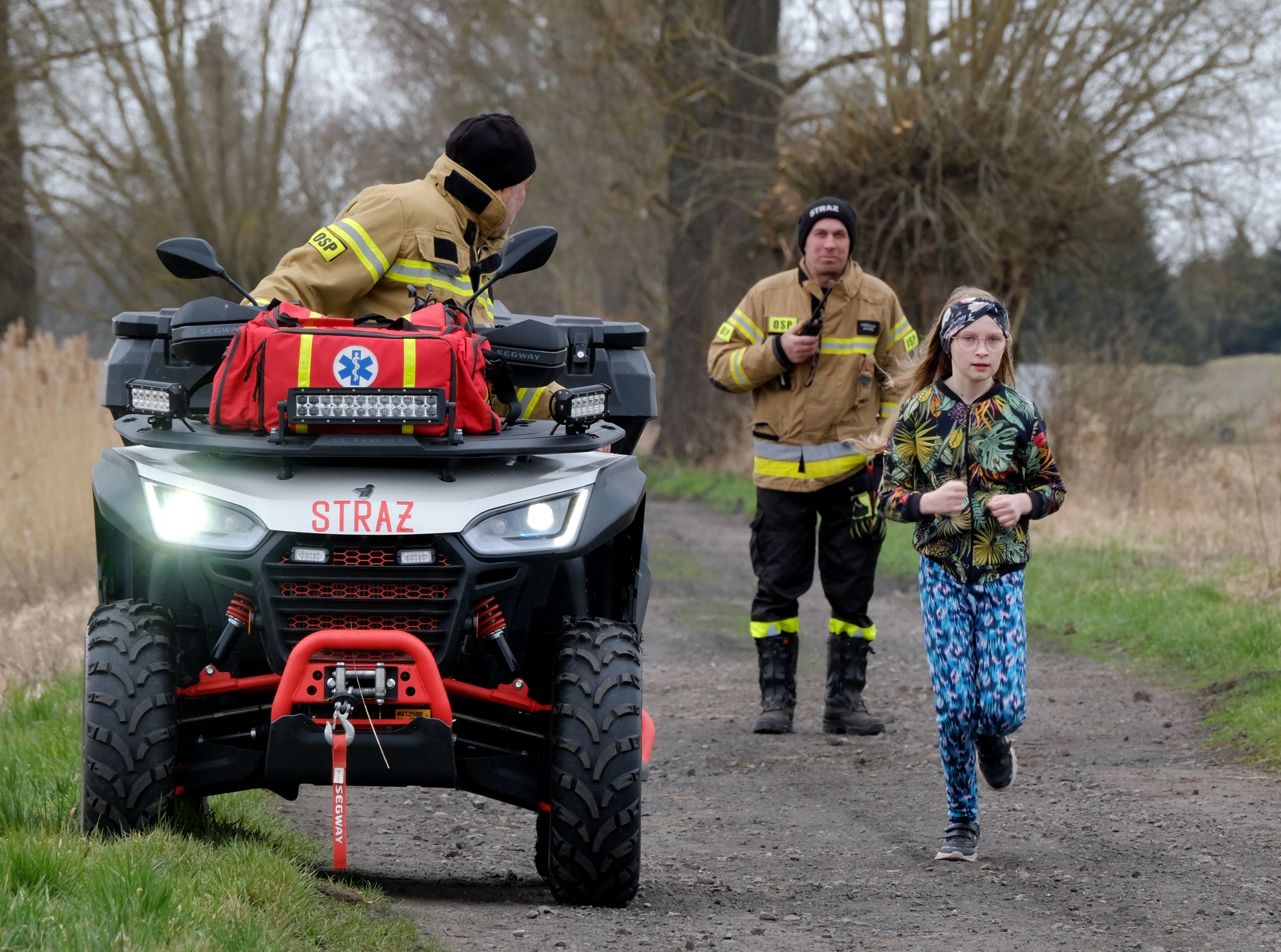 Child jogging on a rural path with rescue personnel and off-road vehicle nearby.