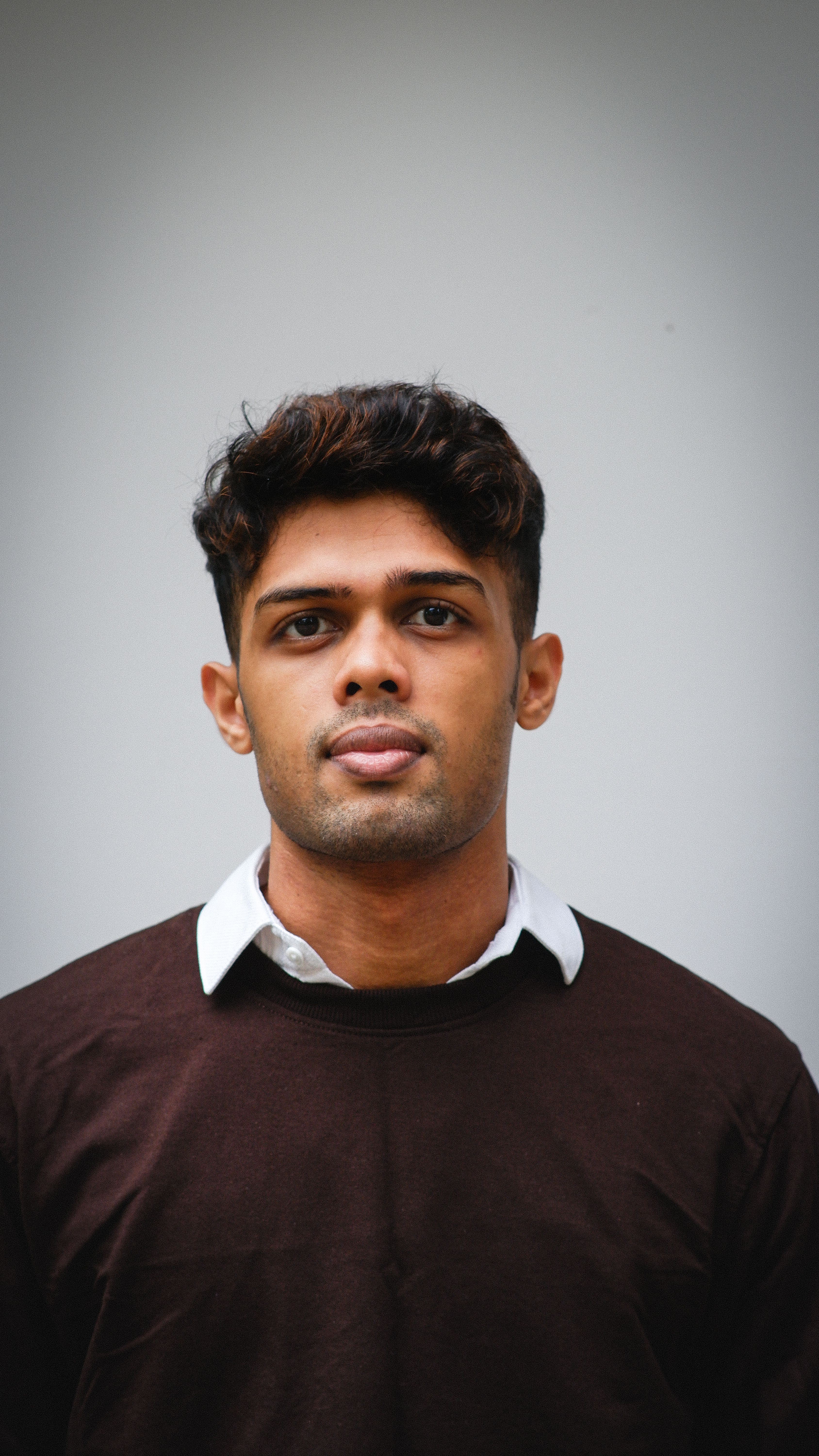 Close-up portrait of a young man with curly hair wearing a brown sweater and white shirt.