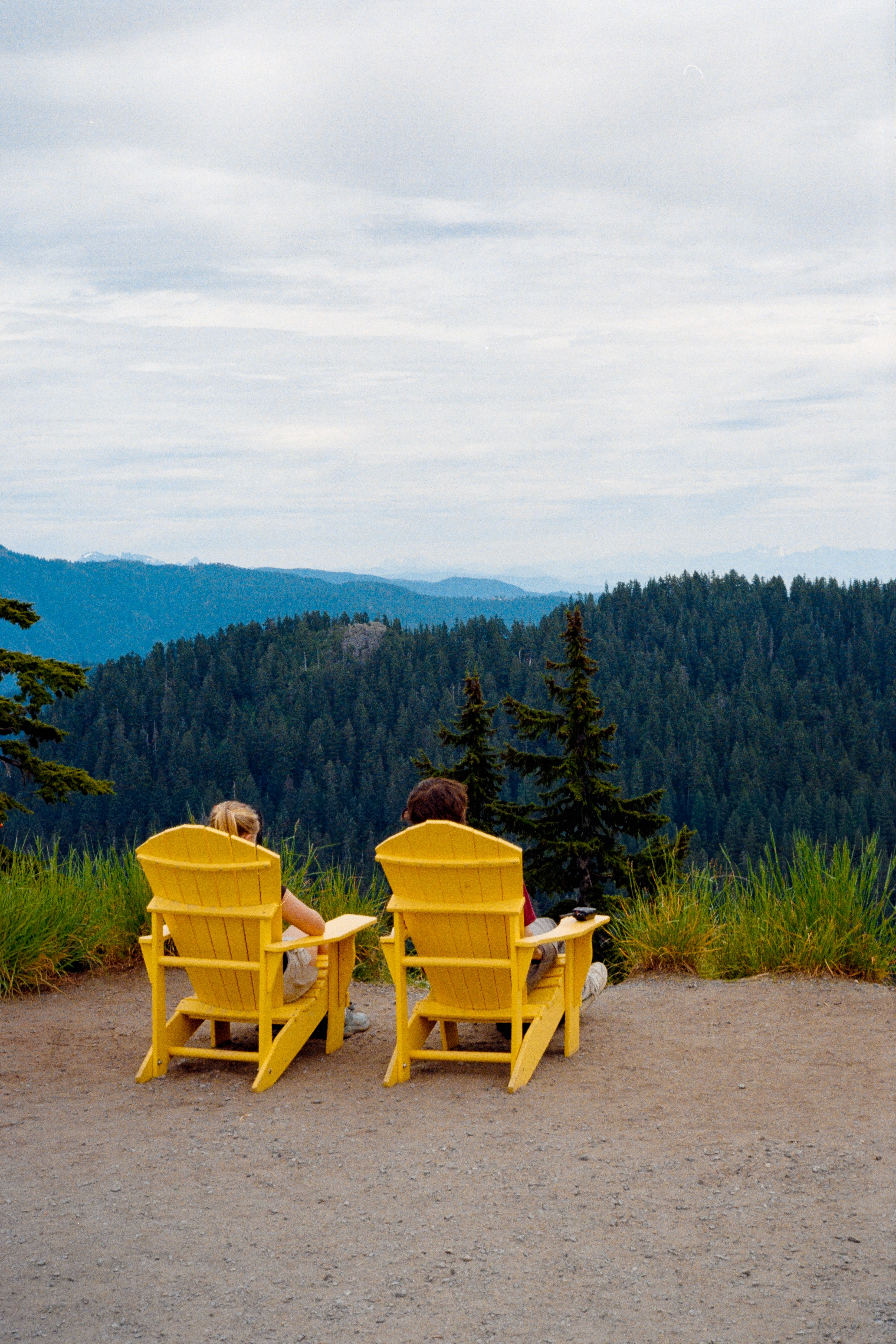 Two people relaxing in yellow Adirondack chairs overlooking a mountain landscape.