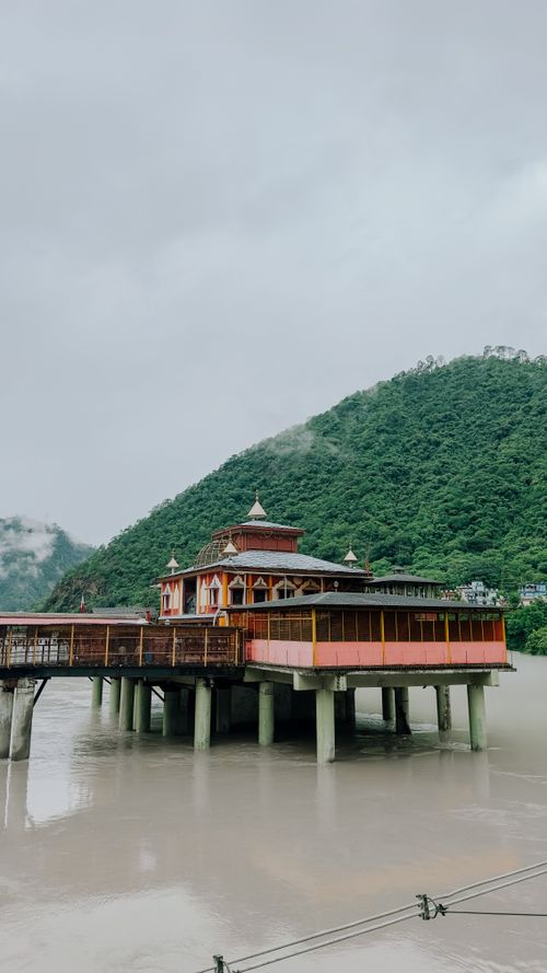 Free A picturesque temple on stilts overlooking a river against a lush green hill backdrop under an overcast sky. Stock Photo