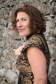 Elegant woman in vintage dress posing by rustic stone wall, day setting.