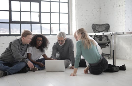 A diverse team of four adults collaborating on a project with a laptop in a bright, modern office setting.