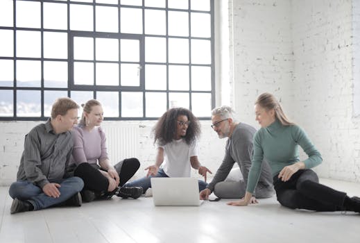 A multicultural group discussing ideas while sitting on the floor with a laptop in a modern office.