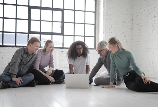 A diverse team of colleagues gathered indoors, enjoying a cheerful and friendly discussion around a laptop.