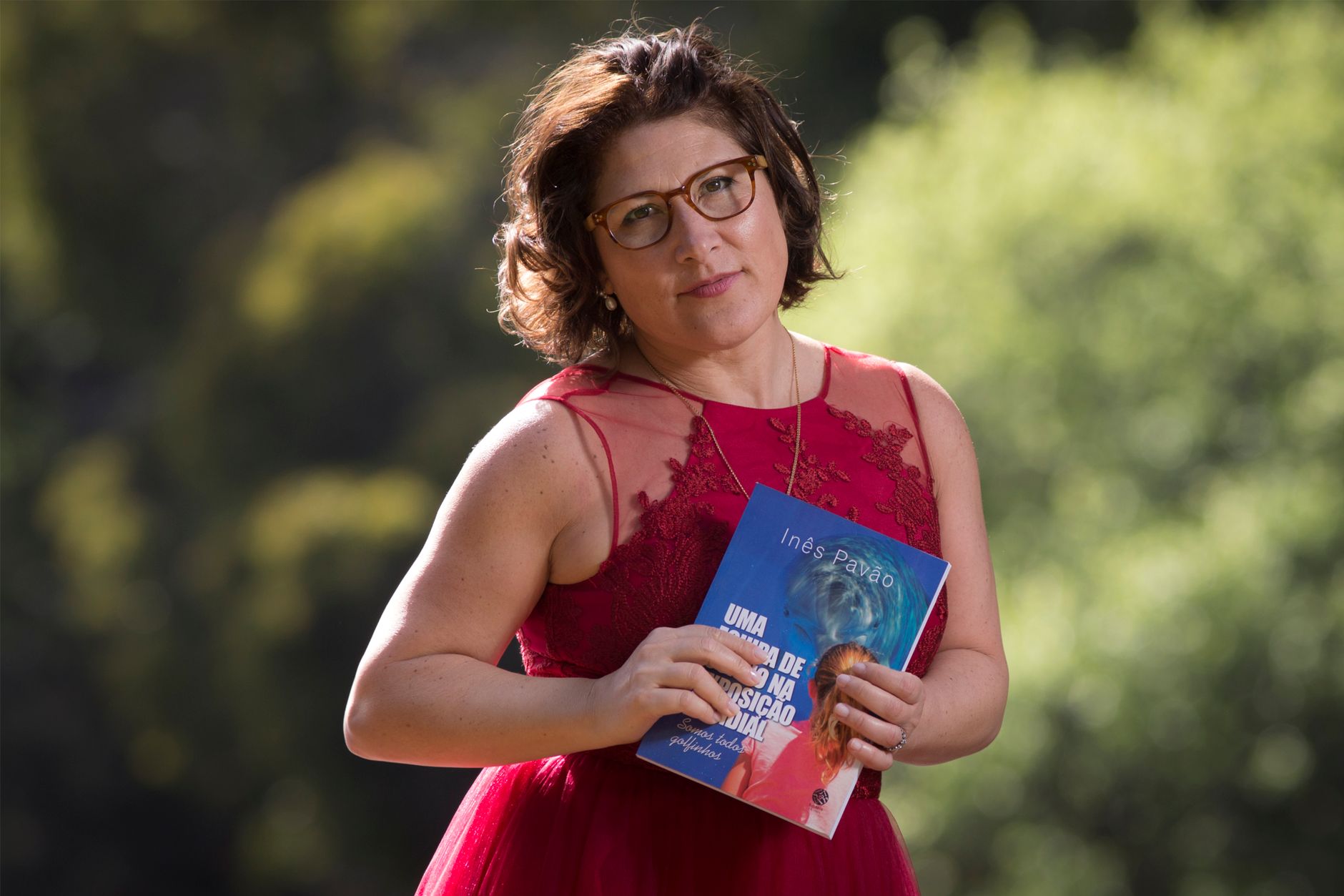 A woman in a red dress holding a book outdoors in a sunny park setting.