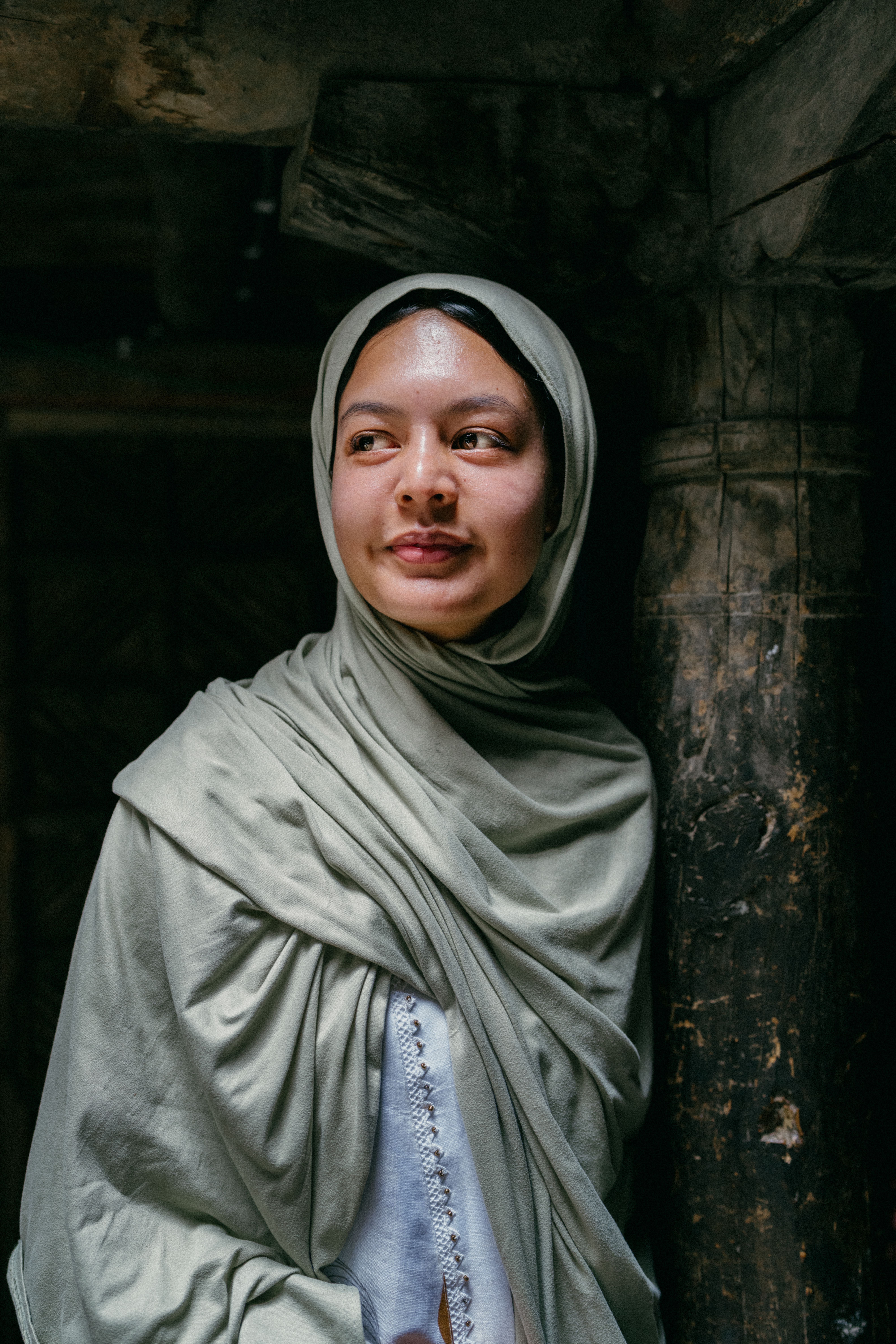 A young woman in soft green hijab, standing indoors, with a thoughtful expression.