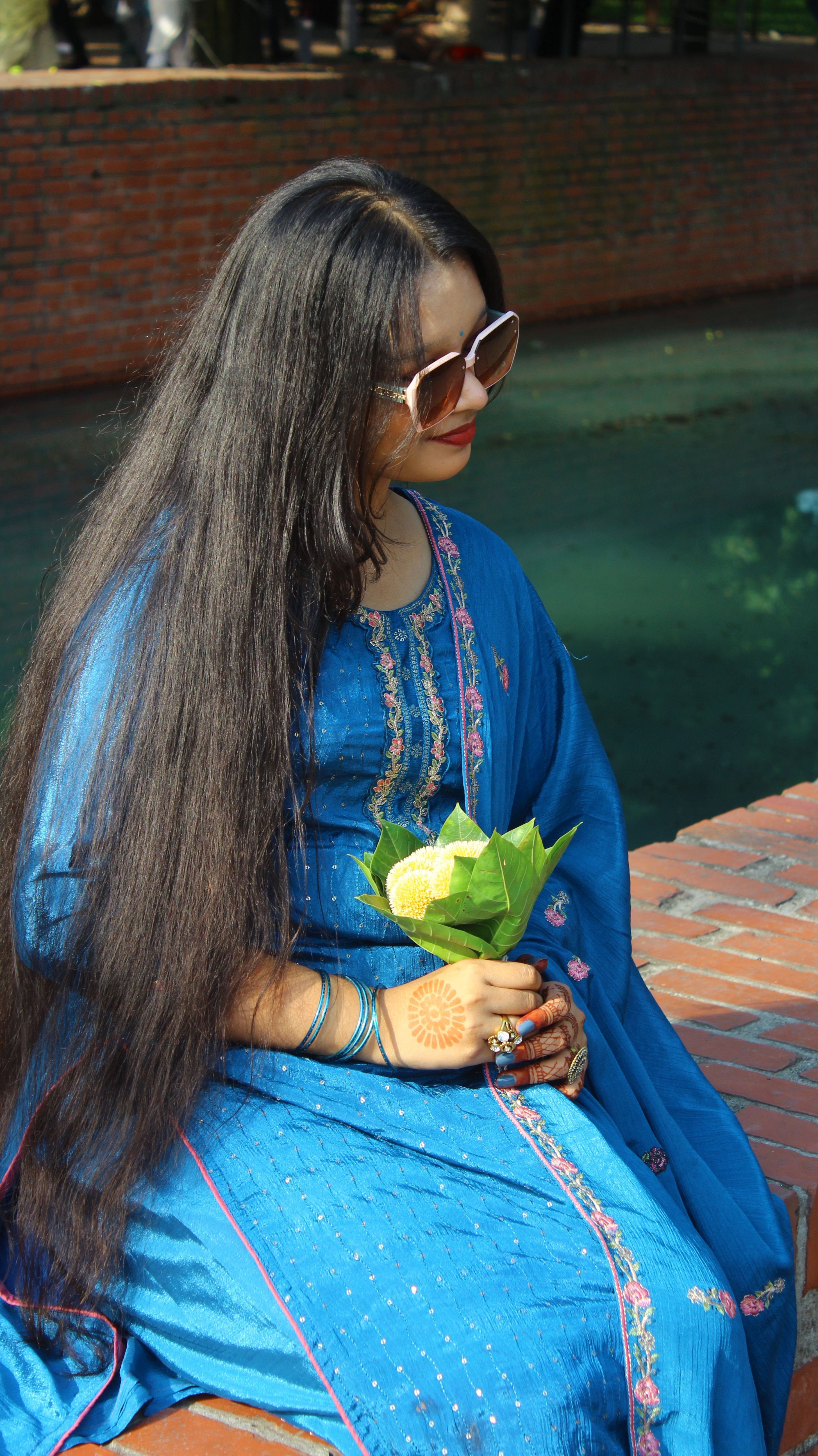 A woman in a blue traditional dress sits outdoors holding flowers, exuding cultural elegance.