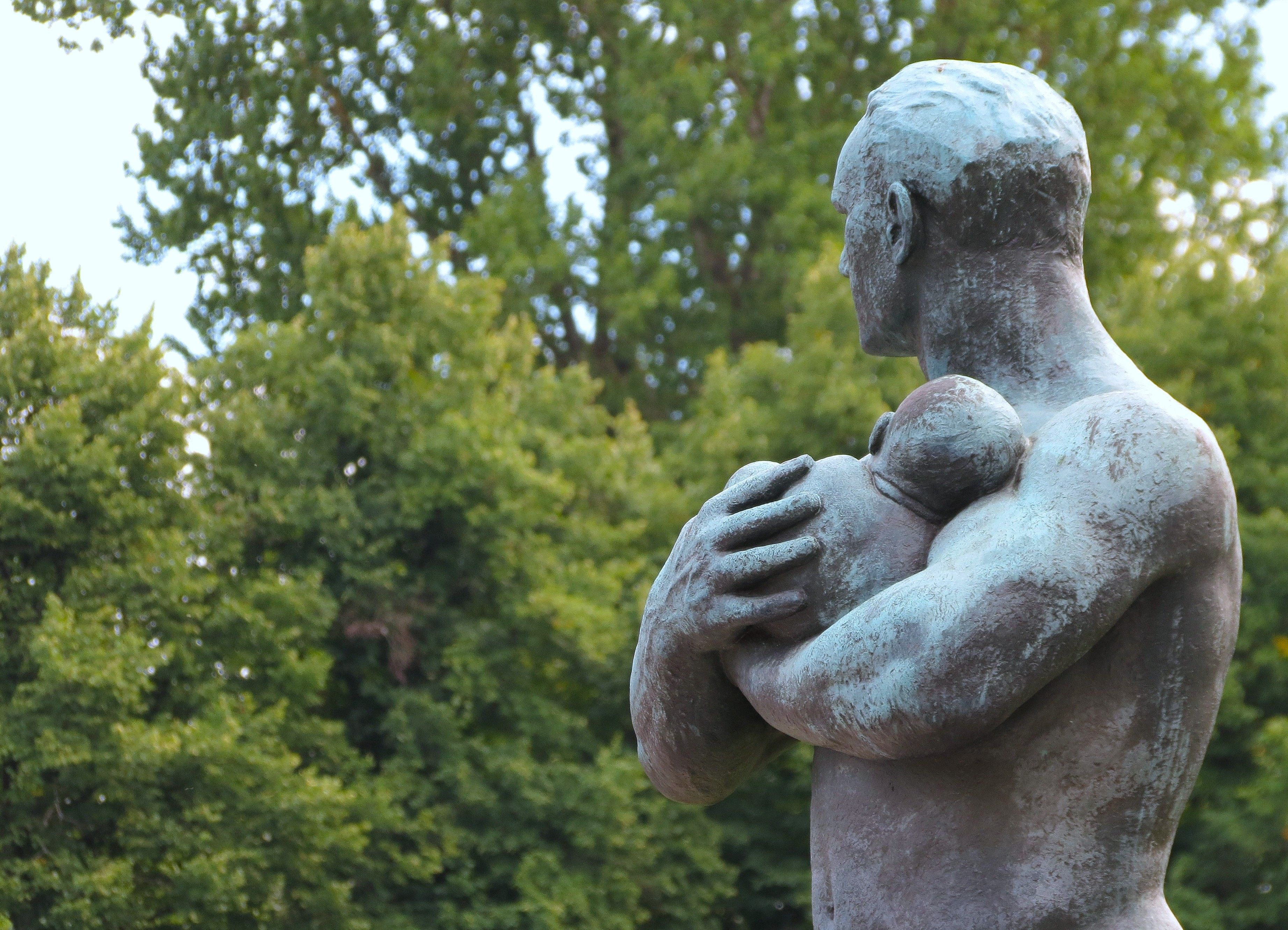 Bronze sculpture depicting fatherly love, set against lush green trees in Oslo park.