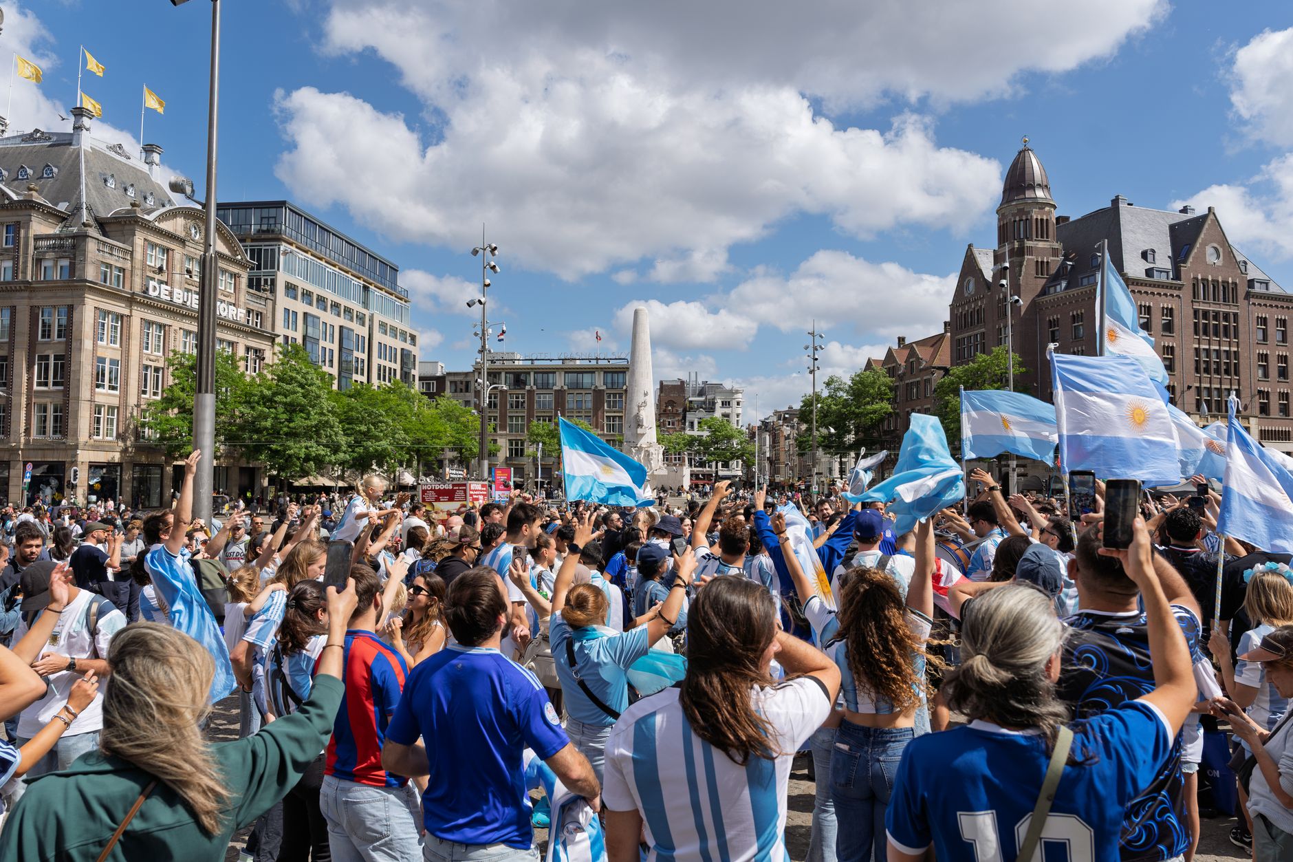 A vibrant crowd celebrates with flags in Amsterdam's Dam Square under a sunny sky.