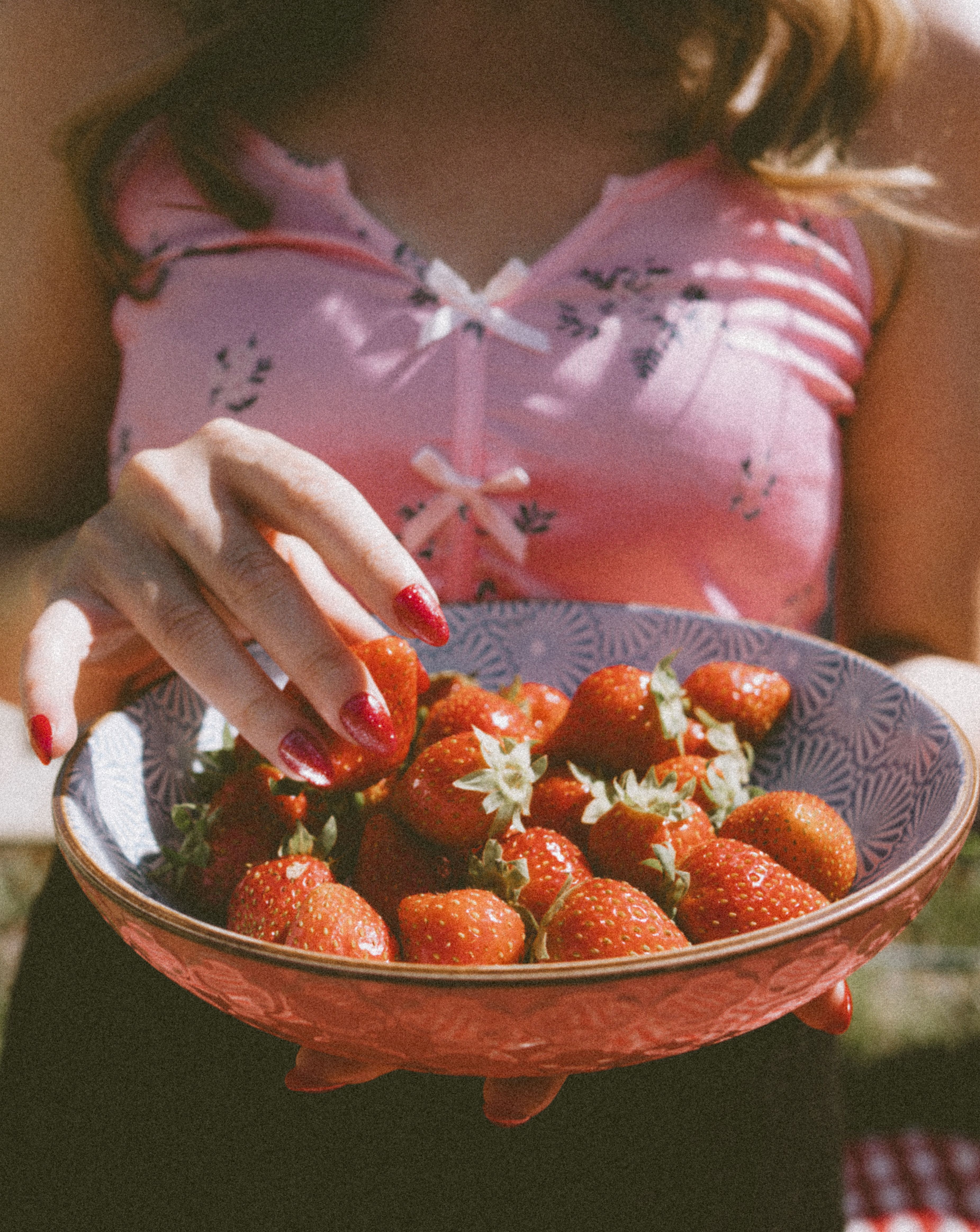 A woman in a floral top holding a decorative bowl filled with ripe strawberries outdoors.