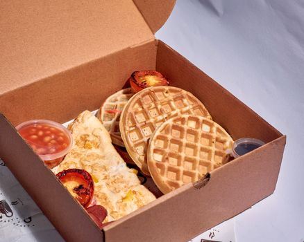 Cardboard takeout box featuring waffles, omelette, and grilled tomatoes, perfect breakfast meal.