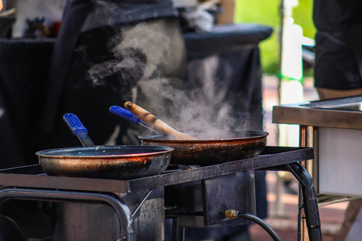 Frying pans with steam on an outdoor stove during a sunny day cooking event.