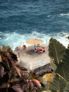 An idyllic seaside spot at La Palma with sunbathers and a straw umbrella near the ocean.