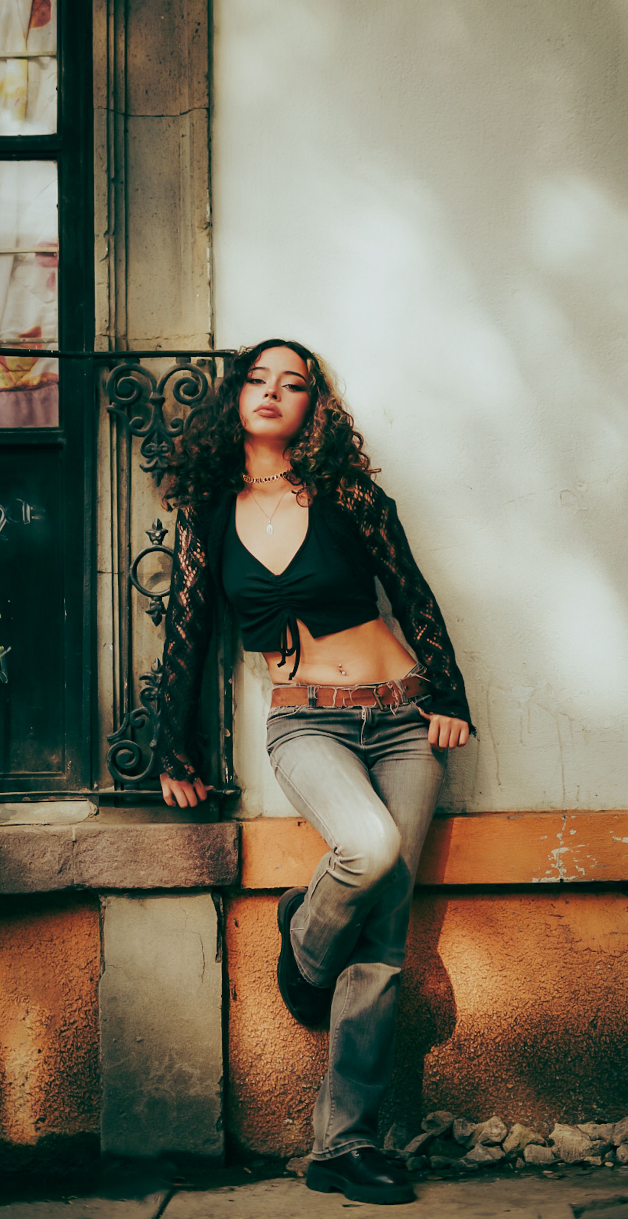 A young woman leans against a wall next to a vintage window, exuding confidence and style.