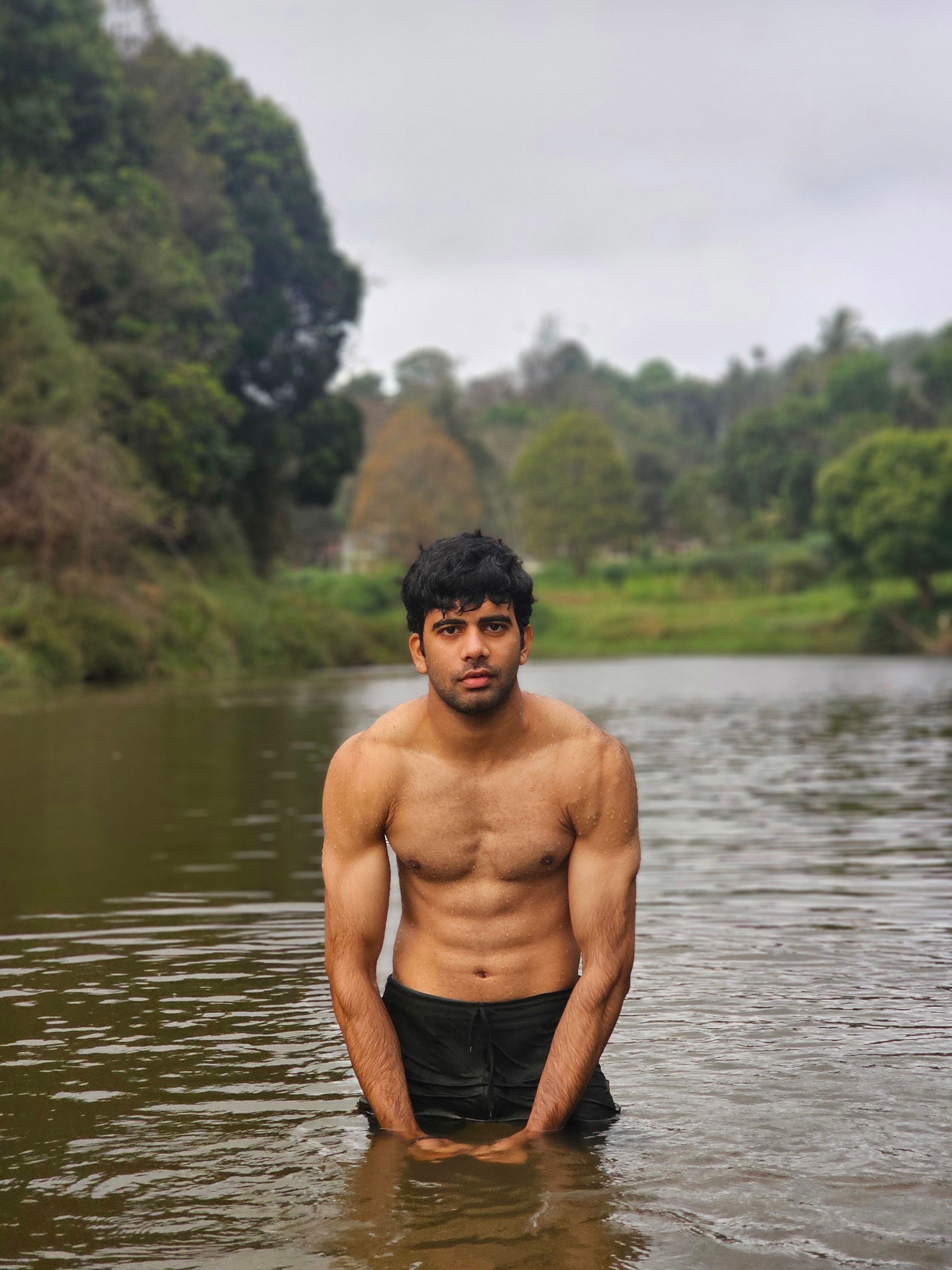 A muscular young man standing in a serene river surrounded by lush greenery, depicting strength and nature.