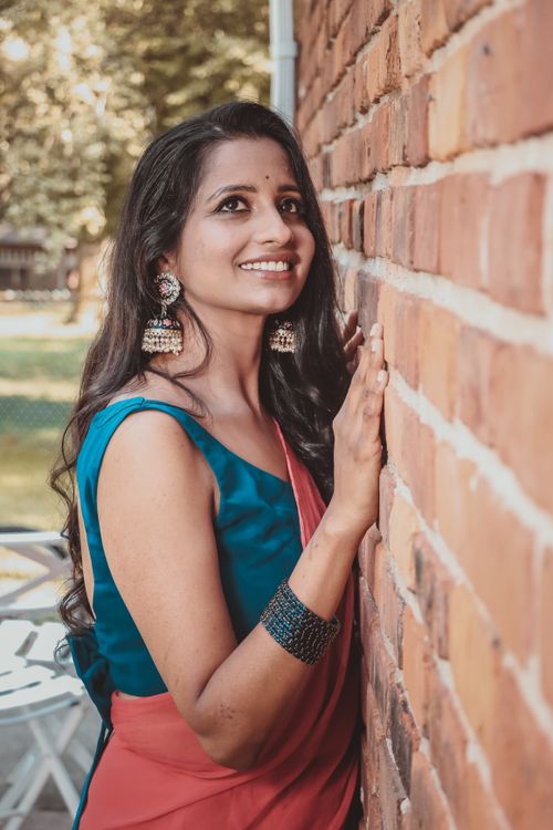 Free Portrait of a smiling woman wearing traditional earrings and a sari, leaning against a brick wall outdoors. Stock Photo