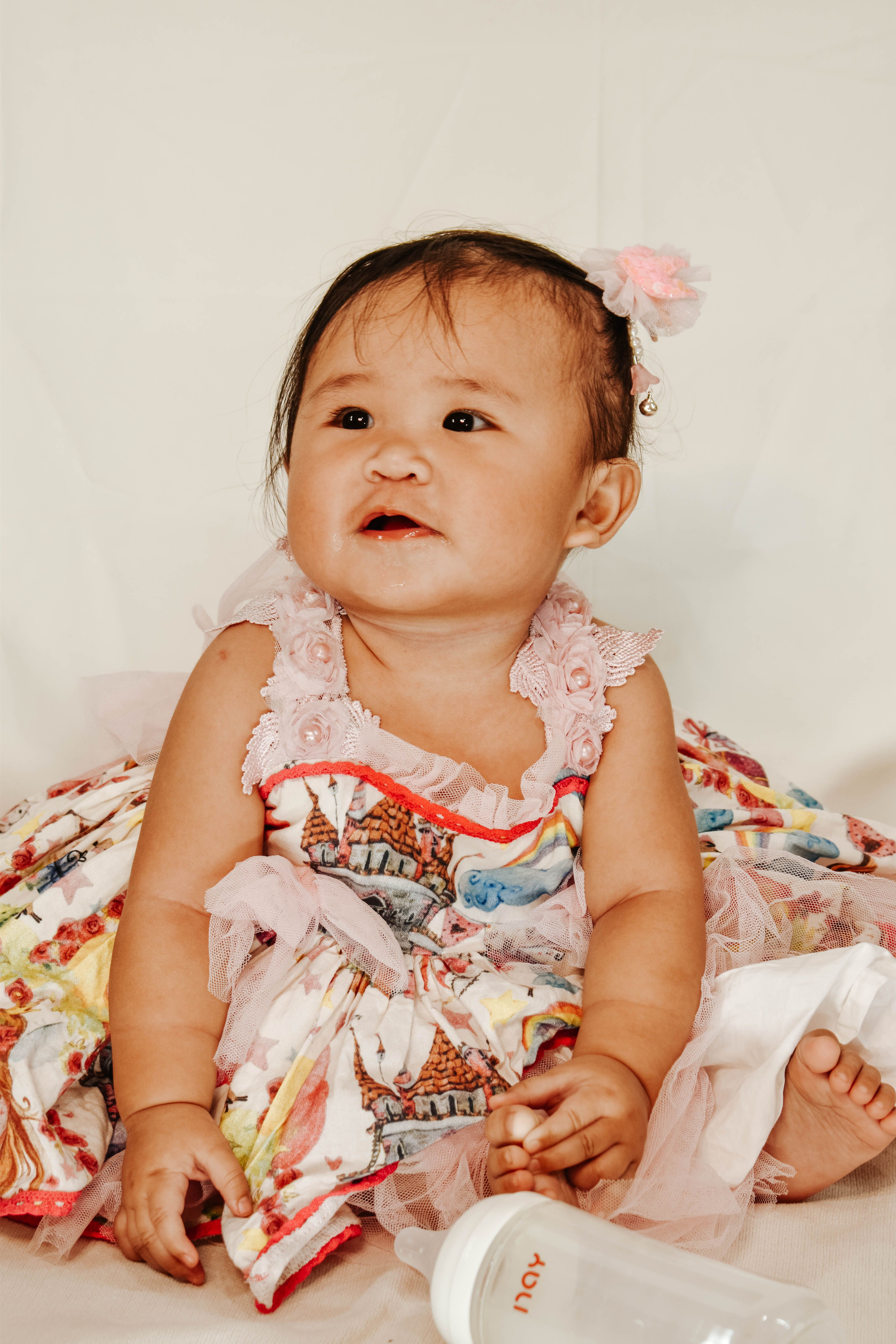 Charming photograph of a baby sitting indoors in a colorful dress with a bottle, showcasing innocence and joyful expression.