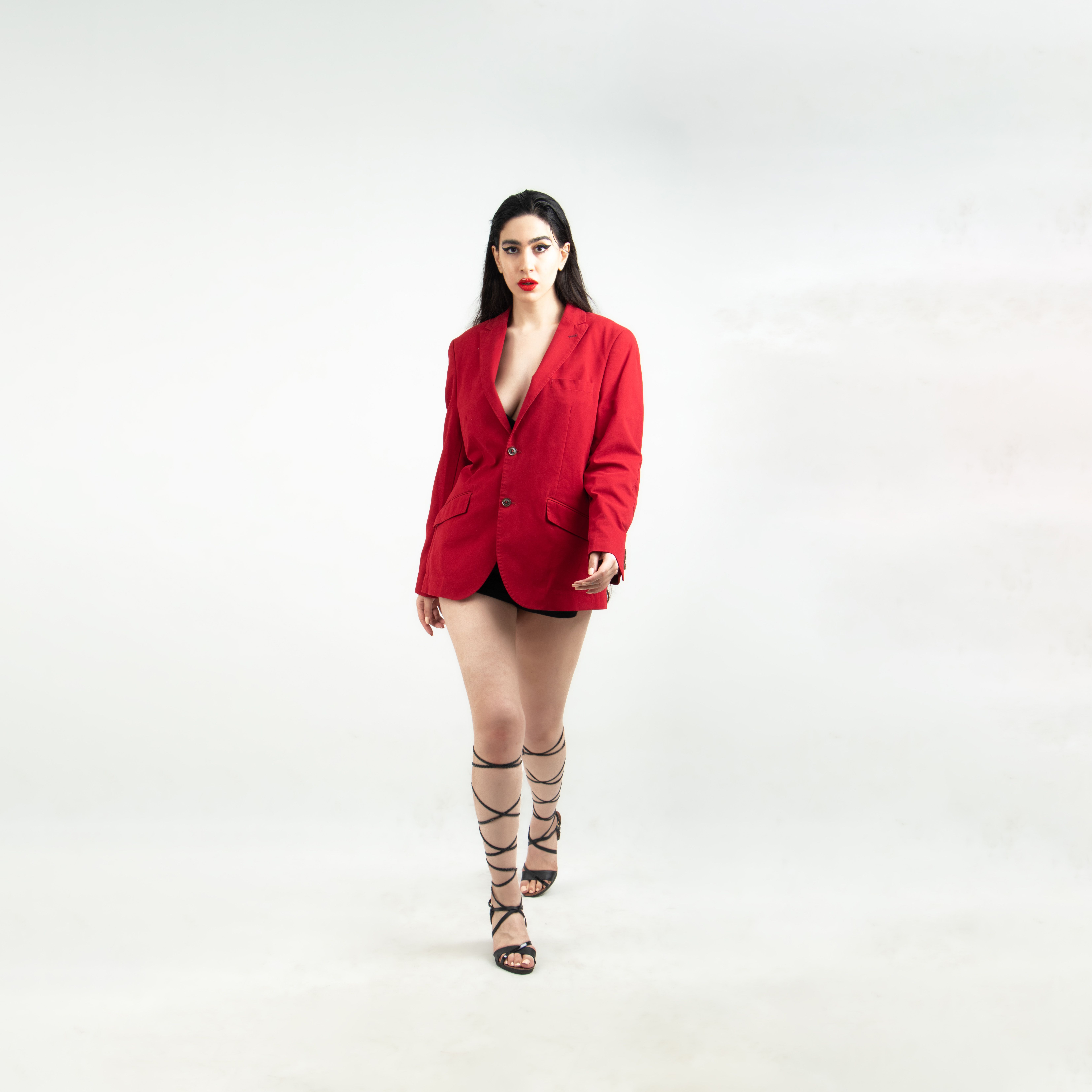 Stylish woman in red blazer and strappy heels posing against a white studio backdrop.