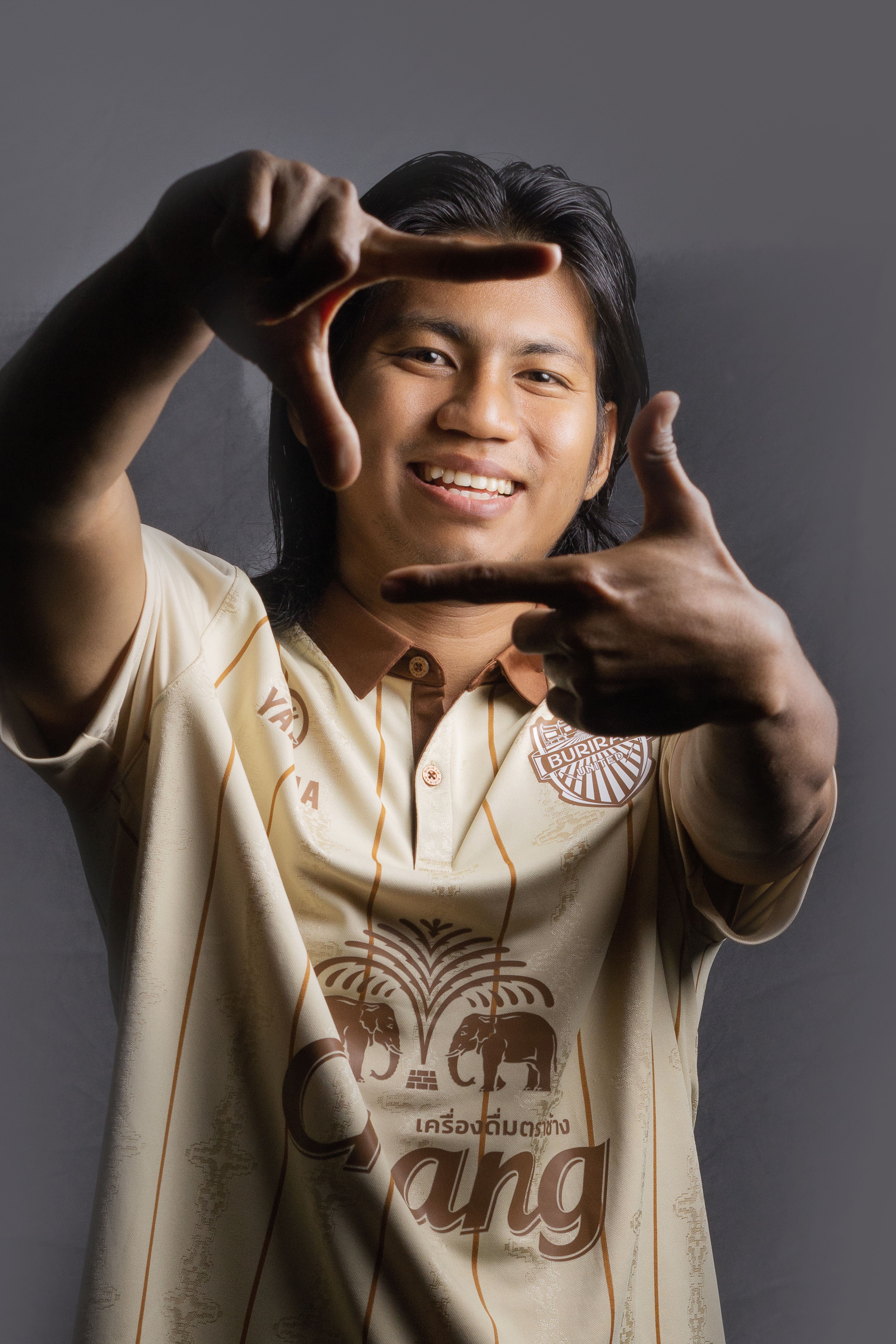 Smiling young man with long hair creating a hand frame gesture wearing a casual shirt.