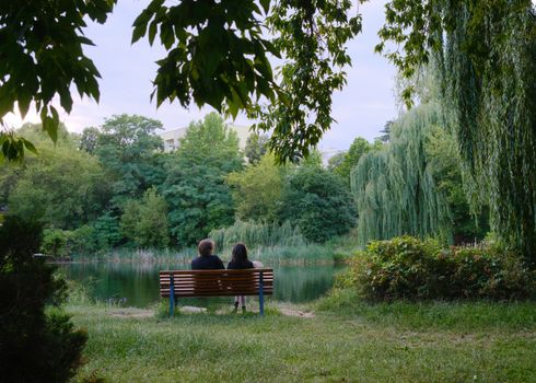 A couple seated on a bench overlooking a serene lake with lush greenery surrounding them.