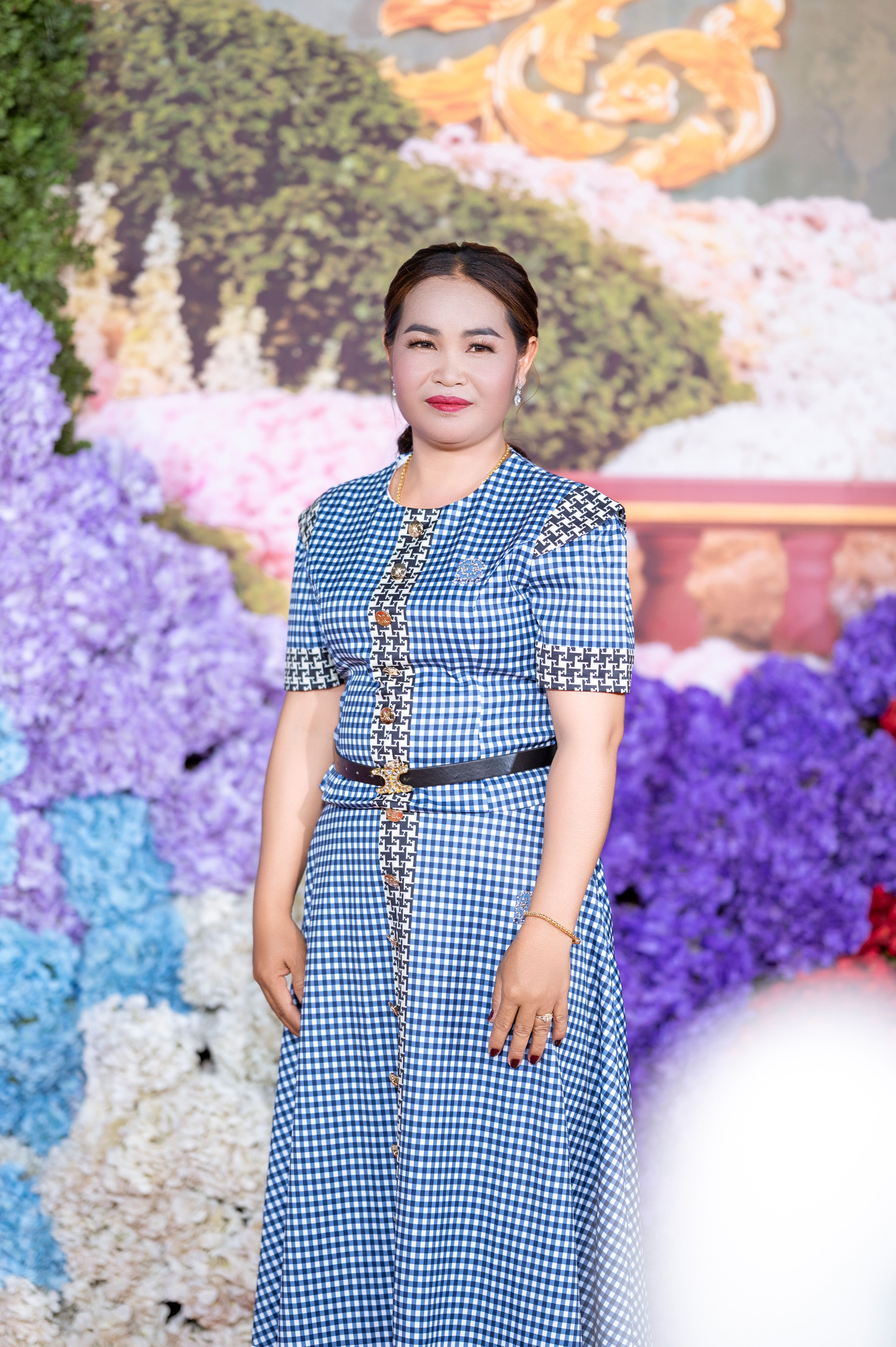 Elegant woman in checkered dress stands against vibrant floral background, Cambodia.