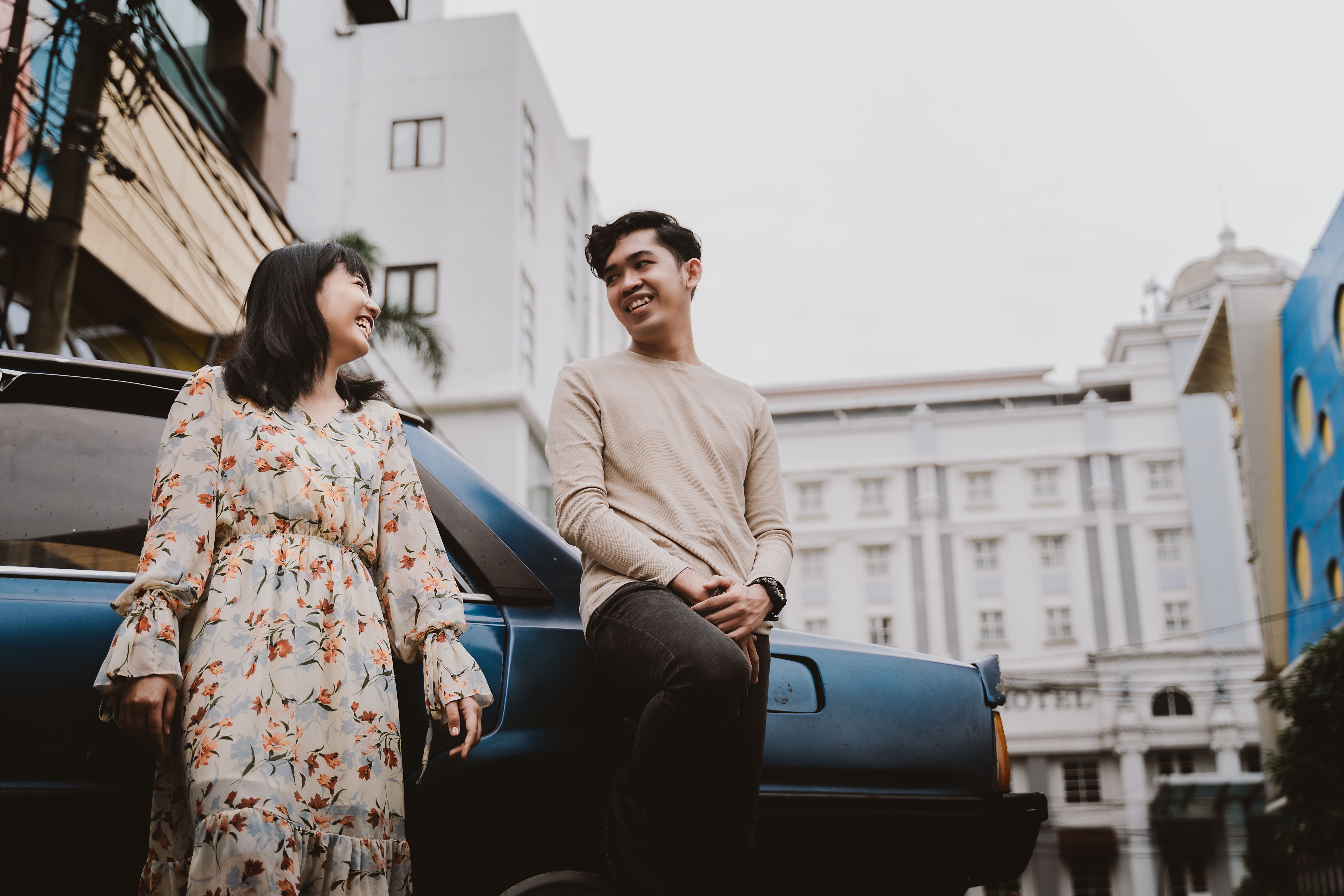 Two young adults laughing beside a classic car with Jakarta architecture in the background.