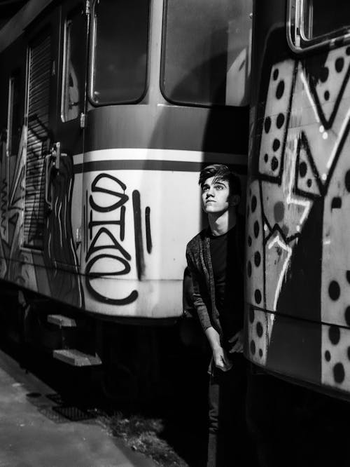 Black and white of pensive young male in casual wear standing between train carriages with graffiti and looking up