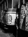 Black and white of pensive young male in casual wear standing between train carriages with graffiti and looking up