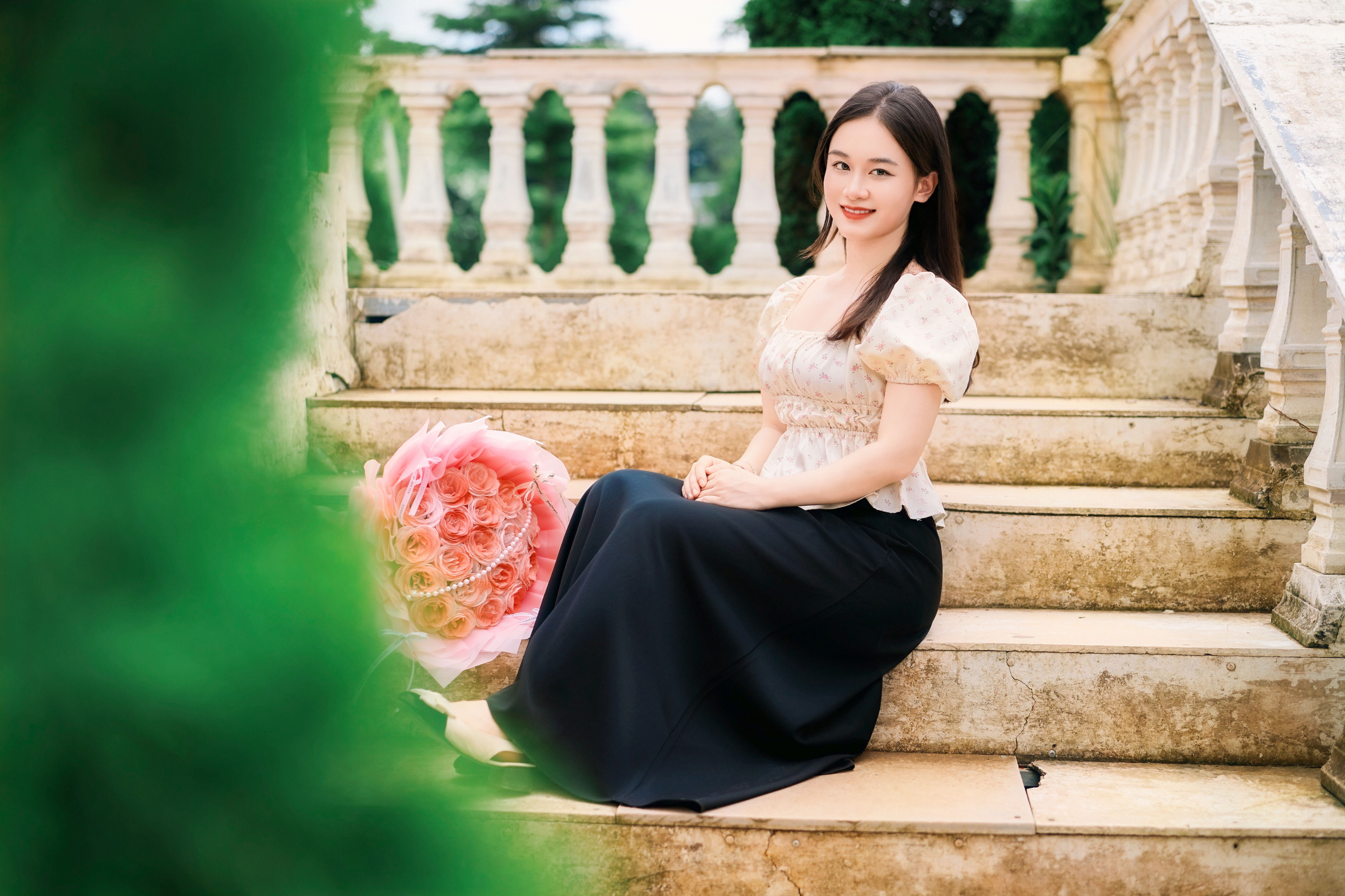 A smiling young woman seated on stone steps outdoors, holding a bouquet of roses.