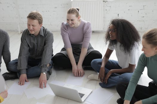 A diverse group of adults engaged in a discussion, sitting on the floor with papers and a laptop.