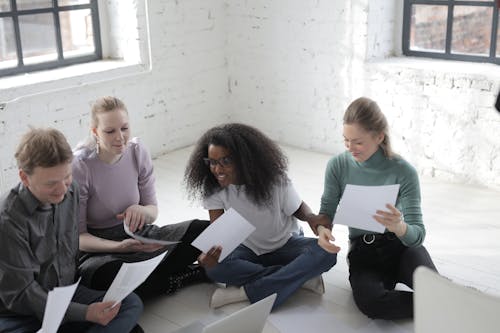 Group of smiling diverse coworkers watching paper while sitting with crossed legs on ceramic floor near open laptop behind brick wall in afternoon