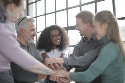 Confident and smiling multiethnic colleagues joining hands and looking at each other while standing near big fenced window in afternoon