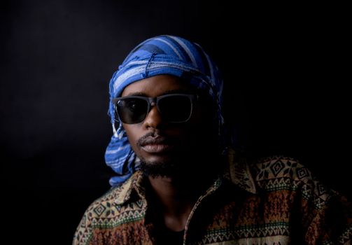 Portrait of a stylish man wearing sunglasses, a blue headscarf, and a patterned shirt against a dark background.