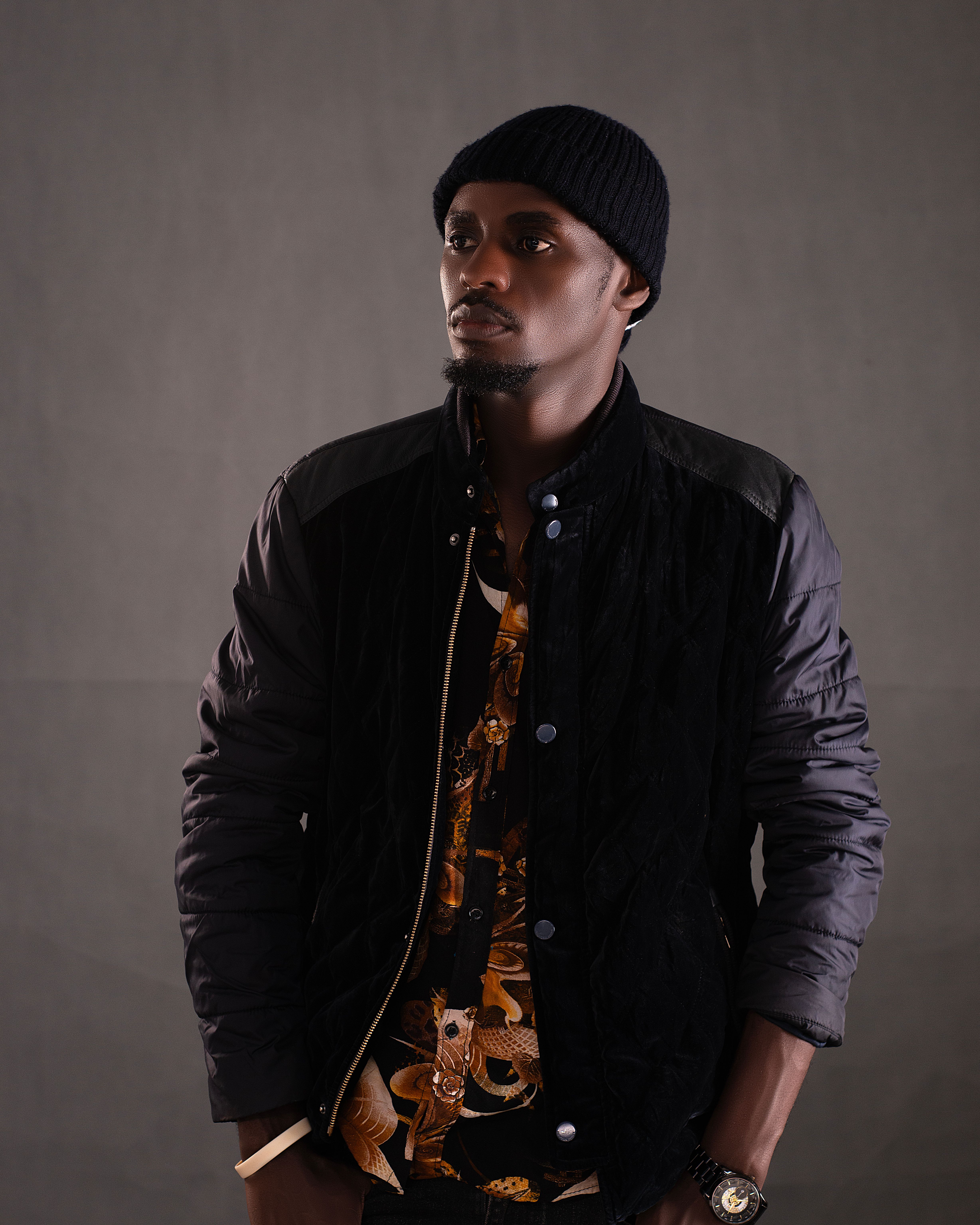 Stylish man in dark jacket and beanie hat posing against a neutral background.