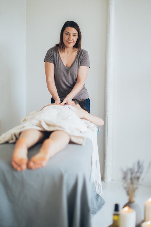 Woman in Gray Crew Neck T-shirt Massaging a Person Lying on Bed