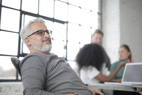 Aged male boss wearing glasses resting in computer chair leaning back and with hands on armpads near desk with laptop while work on project at workplace with industrial interior with group of multiracial coworkers
