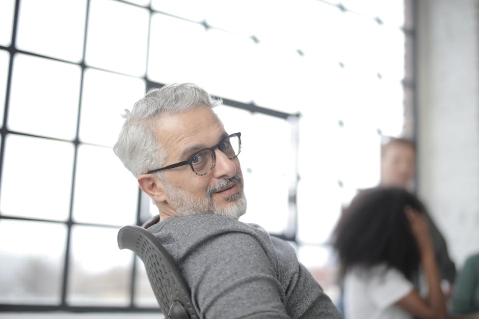 Positive gray haired bearded male manager wearing eyeglasses sitting in computer chair while working in office against big window near group of diverse employees and looking over shoulder