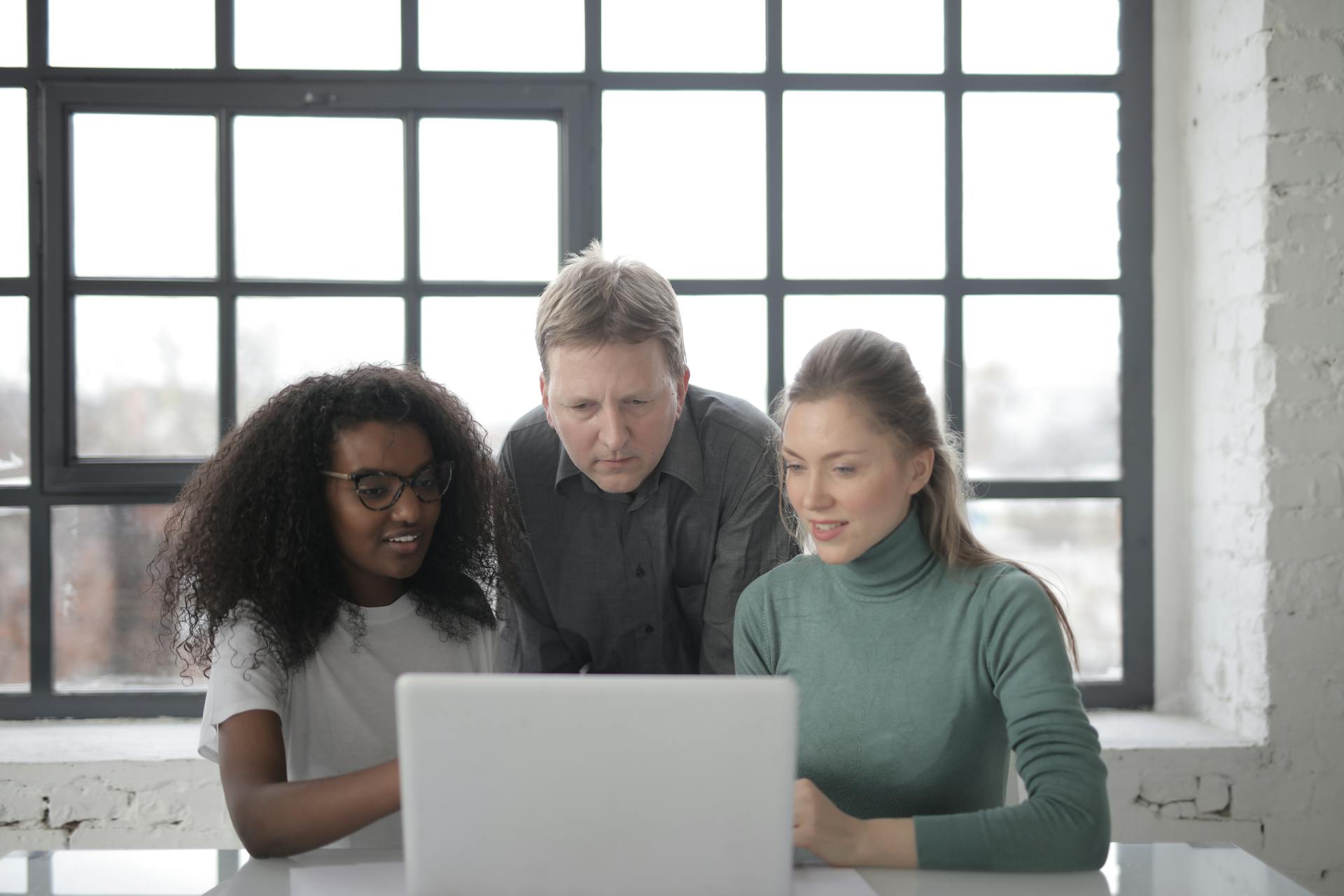 Positive focused multiracial colleagues read information from laptop while teamwork on project in office with industrial interior against big window at daytime