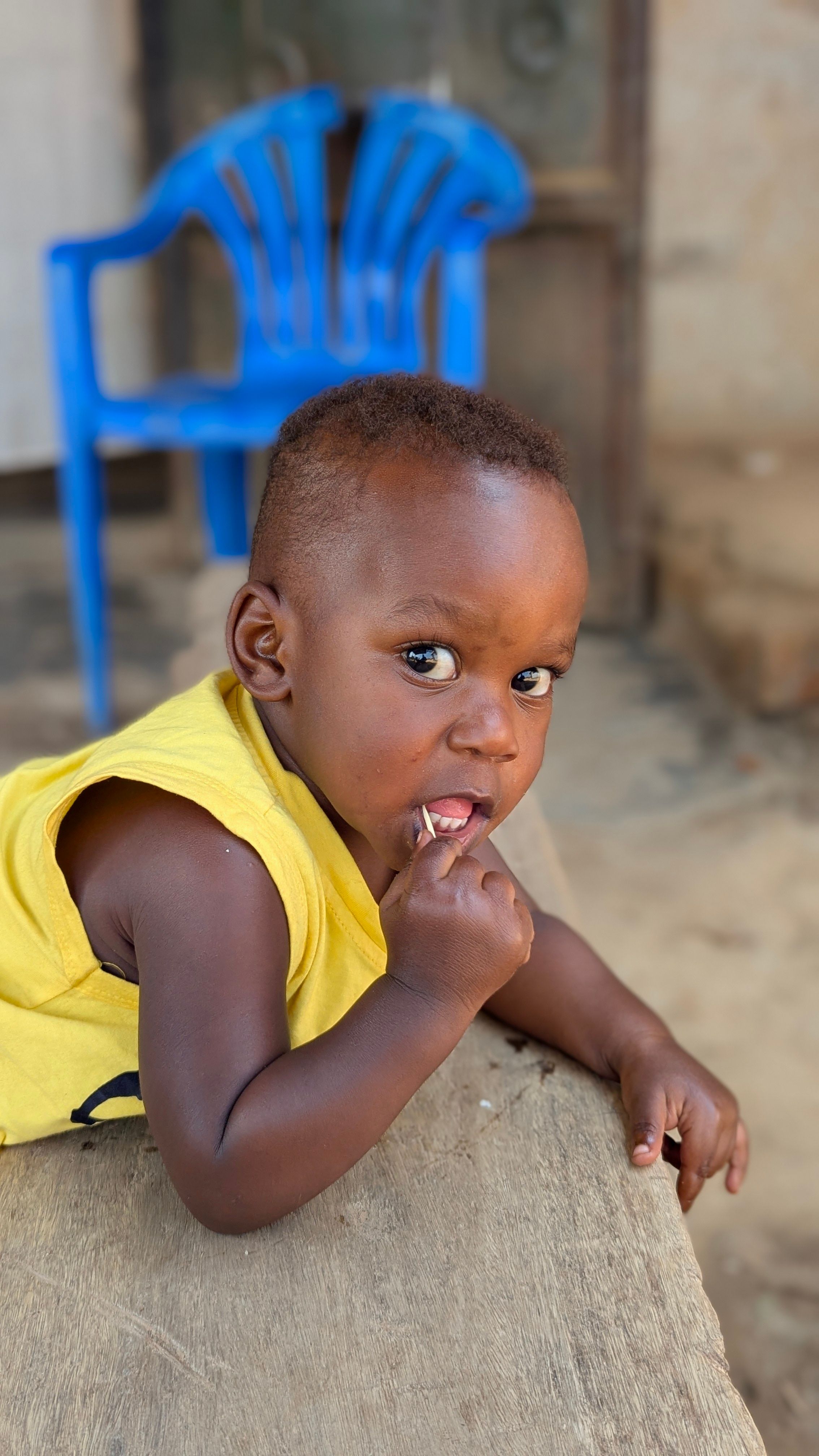 Adorable baby wearing yellow shirt looking curious while chewing on finger outdoors.