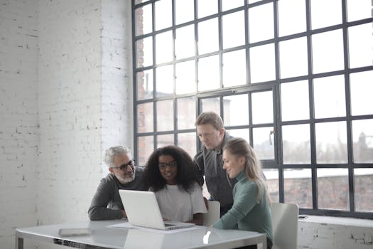 A group of professionals working together on a laptop in a bright, industrial-style office.