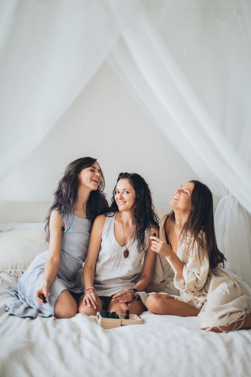 Free Three young women laughing and having fun while sitting on a bed indoors. Stock Photo