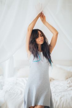 A serene woman stretches with eyes closed in a bright bedroom, exuding calm and relaxation.