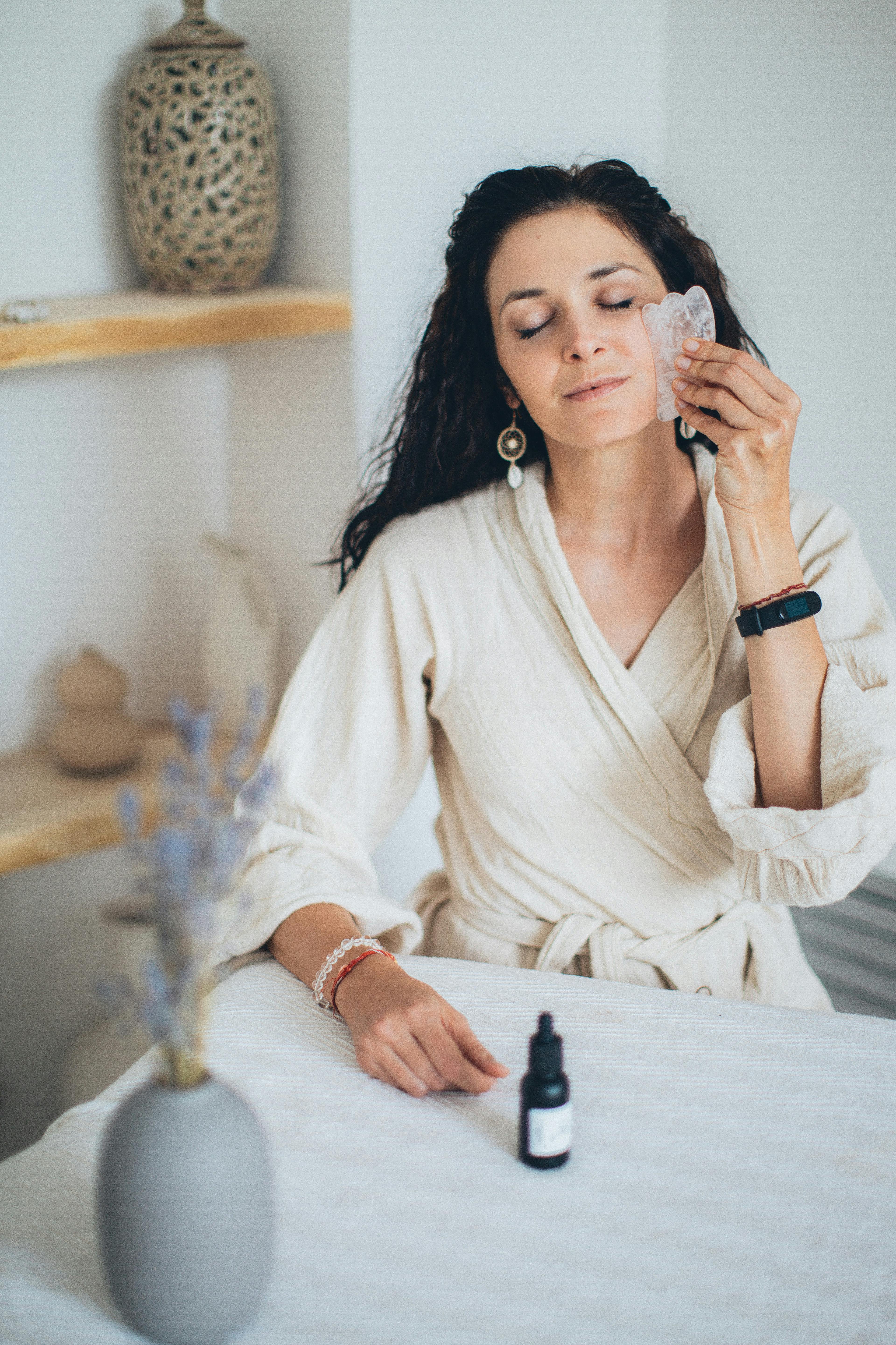 a woman wearing a bathrobe while using a gua sha on her face