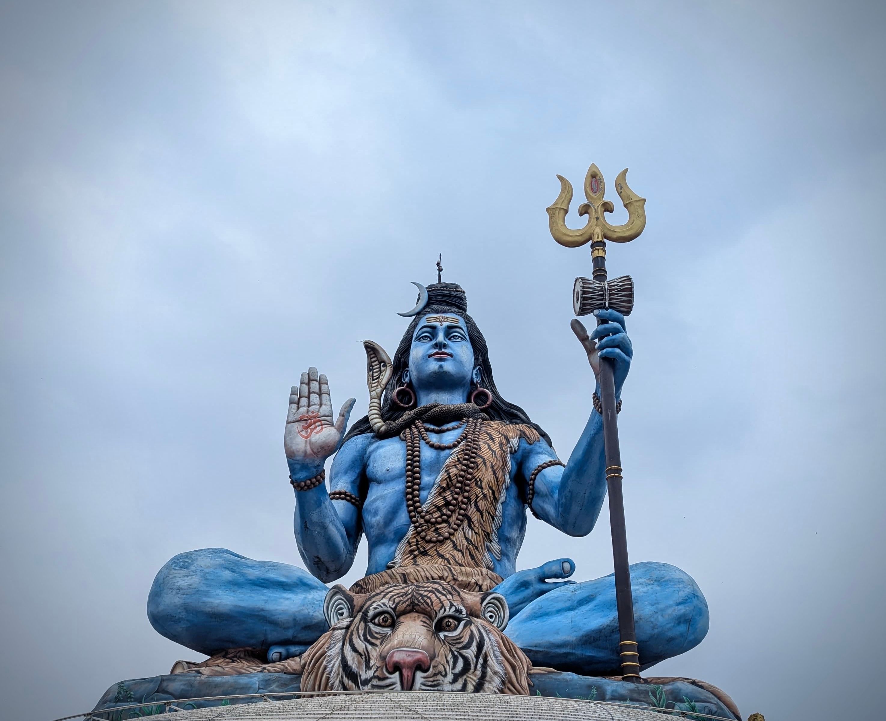 A large blue statue of Hindu deity Shiva holding a trident, set against a cloudy sky.